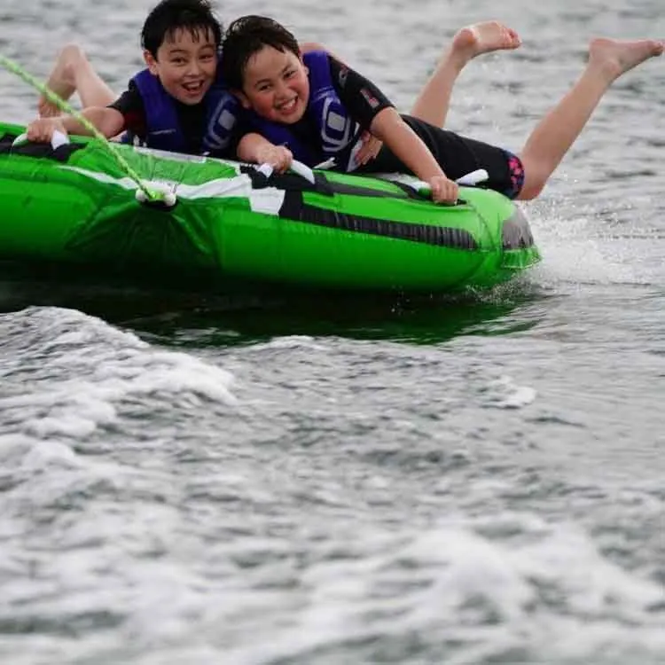Two kids grinning on a green tube behind the boat on Candlewood Lake